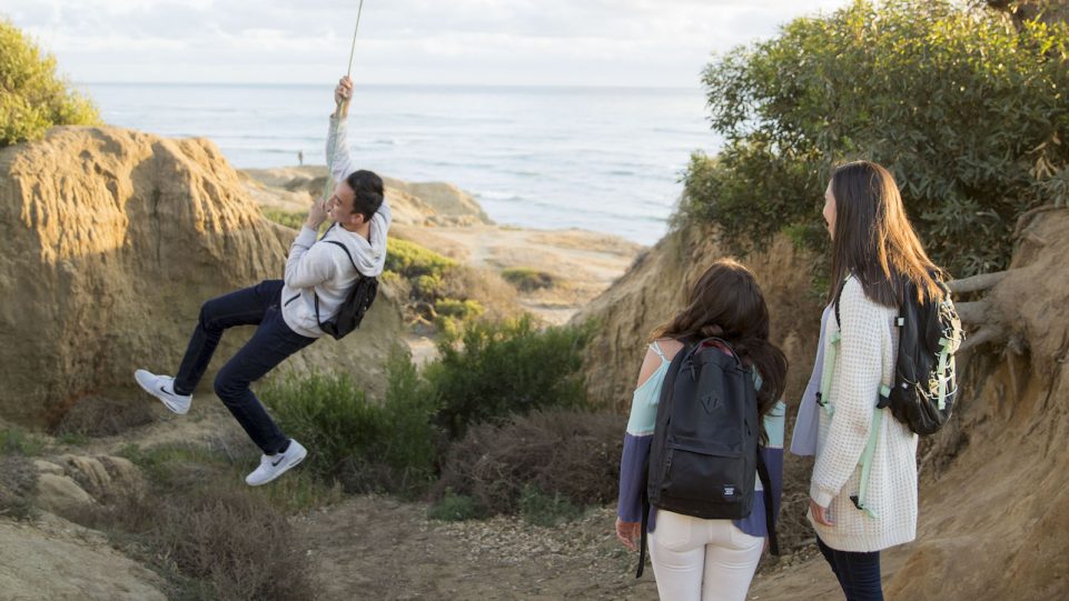 A Male Student Swings on a Rope at Sunset Cliffs Park While Two Female Students Observe